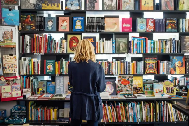 Donna bionda in libreria, circondata da scaffali pieni di libri colorati. Un'atmosfera di lavoro e scoperta tra le pagine.